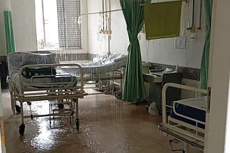 Hospital em João Monlevade com enfermaria masculina parcialmente alagada após vazamento no teto. A imagem mostra leitos hospitalares posicionados lado a lado, com água escorrendo do forro e formando poças no piso. Parte do teto apresenta infiltração visível, e o chão está molhado em diversos pontos, refletindo a iluminação do ambiente.