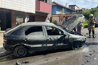 A imagem mostra um carro de passeio completamente queimado, parado ao lado do meio-fio em uma rua residencial durante o dia. O veículo apresenta a lataria escurecida e coberta por fuligem, com sinais claros de incêndio, especialmente na parte dianteira. O capô está aberto, expondo o compartimento do motor, que aparenta ter sido o ponto mais afetado pelas chamas. Nota-se, ainda, a presença de um militar do Corpo de Bombeiros de Monlevade.