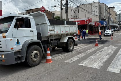 A imagem mostra uma operação tapa-buracos em via urbana, com equipes trabalhando na recuperação do asfalto. Em primeiro plano, há um caminhão basculante branco, estacionado parcialmente sobre a pista, sinalizado com cones de trânsito para organizar o fluxo de veículos. Ao fundo, trabalhadores uniformizados, alguns com coletes refletivos, realizam o reparo do pavimento, espalhando e nivelando o asfalto em um trecho da rua. A área está isolada para garantir a segurança durante o serviço.