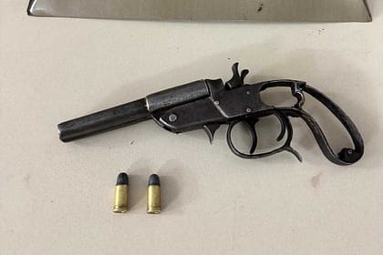 Imagem mostra uma arma de fogo do tipo garrucha apoiada sobre uma superfície clara, acompanhada de duas munições intactas posicionadas à frente. Ao fundo, aparece um suporte metálico em formato triangular com o emblema da Polícia Militar, indicando que o material foi apreendido e apresentado em ambiente institucional. A cena sugere registro oficial de apreensão realizada pelas forças de segurança.