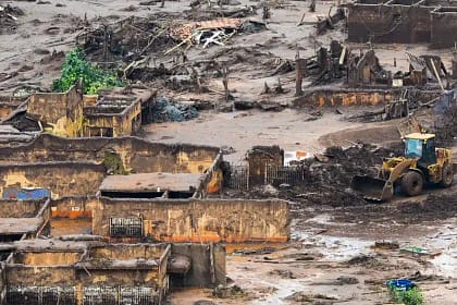 Imagem aérea mostra área devastada pelo rompimento da barragem em Brumadinho. Construções aparecem parcialmente soterradas pela lama, com paredes destruídas e estruturas expostas. O terreno está coberto por rejeitos de mineração, entulho e troncos de árvores.