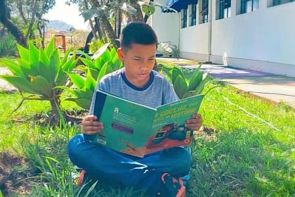 A imagem mostra uma criança sentada sobre a grama, em um ambiente externo e arborizado, lendo um livro infantil. Ela está sentada com as pernas cruzadas, concentrada na leitura, segurando um livro de capa verde com ilustrações coloridas.