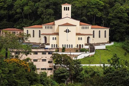 A imagem mostra a Igreja São José Operário em destaque, situada em uma área elevada e cercada por vegetação densa, com morros e árvores ao fundo. O templo possui arquitetura imponente e simétrica, com fachada clara, telhados em tom avermelhado e uma torre central que se eleva acima do conjunto principal. Na parte frontal, observa-se uma grande cruz fixada na fachada, elemento central da composição visual. As laterais apresentam janelas altas e arqueadas, reforçando o estilo religioso tradicional. O acesso à igreja é feito por escadarias e caminhos em níveis, acompanhando o relevo do terreno.