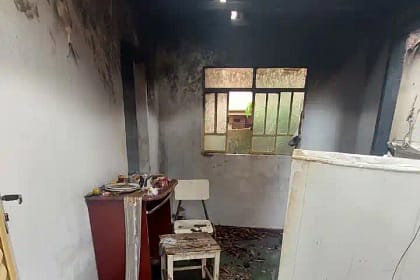 O interior de uma residência aparece severamente danificado por incêndio. As paredes e o teto estão cobertos por fuligem escura, com sinais evidentes de que o fogo se concentrou principalmente na parte superior do cômodo. Há uma abertura no telhado por onde entra luz natural. No ambiente, é possível ver móveis parcialmente queimados, como um pequeno armário, uma cadeira e uma mesa, além de objetos espalhados pelo chão.