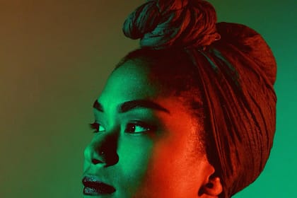 A imagem mostra o retrato de uma mulher vista de perfil, com o rosto levemente voltado para cima e o olhar direcionado para fora do enquadramento. Ela usa um turbante amarrado na cabeça, cobrindo totalmente o cabelo. A iluminação é artística e contrastante, com luz verde incidindo sobre parte do rosto e luz vermelha sobre a outra, criando um efeito dramático. O fundo é liso e desfocado, em tons suaves, destacando a expressão serena e contemplativa da mulher.