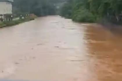 Rio com nível elevado e água barrenta ocupa quase toda a largura do leito, cercado por vegetação densa; ao fundo, céu nublado indica período de chuvas intensas.