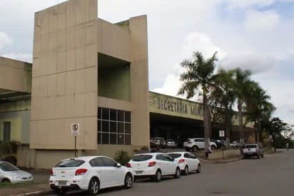 A foto mostra a fachada de um prédio público de grande porte, identificado como Secretaria Municipal de Saúde. O edifício tem arquitetura moderna, com estrutura em tons claros, linhas retas e um bloco vertical destacado na parte frontal. À frente do prédio, há uma via urbana com vários carros estacionados em fila ao longo do meio-fio.
