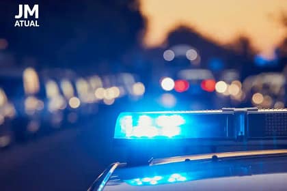 A imagem mostra uma viatura policial em destaque, com o giroflex azul aceso, ocupando a maior parte do enquadramento. O fundo está desfocado, com pontos de luz em tons avermelhados e amarelados, reforçando a ideia de ação policial noturna ou em ambiente urbano.