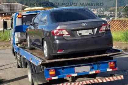 Um automóvel sedan de cor cinza escuro está sendo transportado sobre a plataforma de um caminhão-guincho azul, em via urbana. O veículo aparece visto pela traseira, com a placa parcialmente desfocada. Ao fundo, é possível observar casas, vegetação e uma área urbana em região montanhosa.