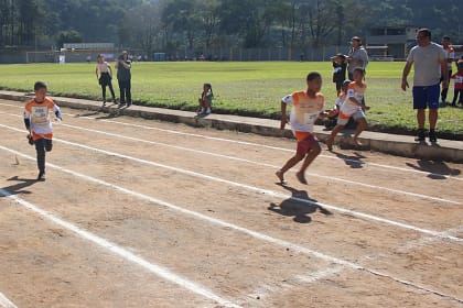 A imagem retrata uma prova de atletismo infantil, realizada em uma pista de terra demarcada, onde crianças disputam uma corrida em clima de competição e integração. Os atletas, usando camisetas padronizadas com numeração, correm descalços ou com calçados leves.