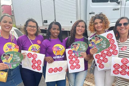 A imagem mostra seis mulheres reunidas ao ar livre, sorrindo para a câmera e segurando materiais de campanha. Elas vestem camisetas roxas com ilustrações e mensagens de conscientização sobre a proteção de crianças e adolescentes. Cada uma segura folhas com vários adesivos vermelhos e leques informativos com o slogan “Pule, brinque e cuide”, relacionados à campanha de combate à exploração sexual infantil.