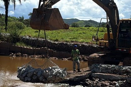 A imagem mostra uma obra de reforço em um curso d’água, em uma barragem ou dique. Uma escavadeira hidráulica, posicionada à margem do rio, utiliza a concha para içar uma rede metálica cheia de pedras, conhecida como gabião, que será utilizada na contenção da estrutura. Ao lado da máquina, um trabalhador acompanha a operação em pé sobre o terreno de terra e pedras.