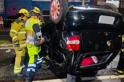 A imagem mostra um veículo preto capotado após acidente em Monlevade, com as rodas voltadas para cima, imobilizado no meio de uma via urbana durante a noite. O automóvel apresenta danos significativos na lataria, especialmente na parte traseira e lateral, indicando a força do impacto. Ao lado do carro, dois militares do Corpo de Bombeiros, devidamente equipados com capacetes, lanternas e uniformes de proteção, atuam no atendimento à ocorrência.