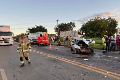 A imagem mostra a cena de um acidente na BR-381, com um carro de passeio fortemente danificado após uma colisão frontal. O veículo aparece com a parte dianteira completamente destruída e parado sobre a pista, cercado por destroços. No local, há atuação de equipes de resgate e segurança. Um bombeiro caminha pela rodovia, enquanto uma viatura do Corpo de Bombeiros está parada mais à frente, com o giroflex ligado. Cones foram posicionados para sinalizar o trecho e organizar o trânsito. Ao fundo, é possível ver caminhões, outros veículos parados e um grupo de pessoas observando a ocorrência às margens da rodovia. O cenário indica atendimento emergencial e interdição parcial da pista após o impacto.