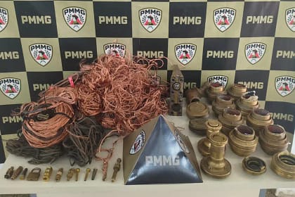 A imagem mostra diversos materiais apreendidos expostos sobre uma mesa, tendo ao fundo um painel institucional com a identificação da PMMG. Em destaque, há grande quantidade de fios e cabos de cobre enrolados e soltos, além de conexões metálicas, peças industriais de latão, registros, bicos e outros componentes utilizados em sistemas industriais. Os itens estão organizados lado a lado para registro da ocorrência, com um suporte triangular da corporação posicionado ao centro da mesa. A foto registra o material recuperado em ação da Polícia de Monlevade, evidenciando a apreensão dos objetos e a atuação policial no combate à receptação no município.