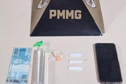 A imagem mostra materiais apreendidos pela Polícia Militar, dispostos sobre uma superfície clara, com um suporte ao fundo exibindo a sigla “PMMG”. Entre os itens estão uma cédula de R$ 100, um frasco plástico contendo lança-perfume, três papelotes de substância semelhante à cocaína, um comprimido de ecstasy e um aparelho celular. Os objetos estão organizados lado a lado, compondo o registro da ocorrência policial. Notícias de nova era