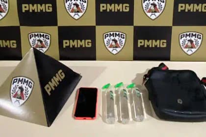 A imagem mostra os materiais apreendidos pela Polícia Militar durante a ocorrência registrada em Nova Era. Sobre uma mesa branca, estão expostos três frascos de lança-perfume, um aparelho celular com capa vermelha e uma bolsa preta tipo pochete. Ao lado dos objetos, aparece um suporte triangular com a sigla da corporação, posicionado diante de um painel ao fundo com o padrão xadrez nas cores preta e amarela, estampado com o emblema da PMMG. A foto integra as notícias de Nova Era relacionadas à prisão de dois suspeitos por tráfico de drogas durante evento carnavalesco no Centro da cidade.