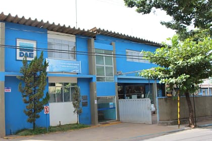 A imagem mostra a sede do Departamento de Águas e Esgotos (DAE) em um prédio de dois andares, pintado em tons de azul e branco. Na fachada, há o logotipo do DAE e uma placa com mensagem institucional sobre o uso consciente da água.