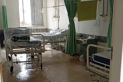 Hospital em João Monlevade com enfermaria masculina parcialmente alagada após vazamento no teto. A imagem mostra leitos hospitalares posicionados lado a lado, com água escorrendo do forro e formando poças no piso. Parte do teto apresenta infiltração visível, e o chão está molhado em diversos pontos, refletindo a iluminação do ambiente.