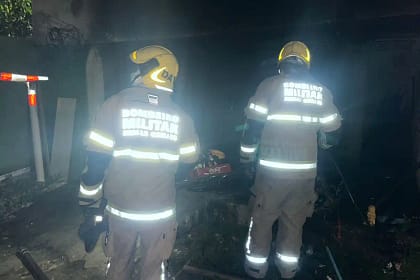 A imagem mostra bombeiros em João Monlevade atuando durante a noite em uma ocorrência. Dois militares do Corpo de Bombeiros aparecem de costas, utilizando capacetes amarelos e uniformes com faixas refletivas, que brilham sob a iluminação do local. O ambiente está escuro e com presença de fumaça, sugerindo atendimento a um incêndio recente. No chão, é possível observar equipamentos e materiais próximos a uma área aparentemente queimada. A cena transmite a atuação da equipe no trabalho de rescaldo e verificação da área atingida.