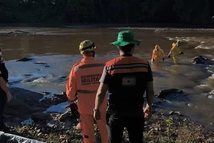 Em Nova Era, equipes do Corpo de Bombeiros e da polícia acompanham uma operação de resgate em um trecho do Rio Piracicaba. Na imagem, militares uniformizados estão próximos às margens e dentro da água, entre rochas, realizando o trabalho de recuperação do corpo.