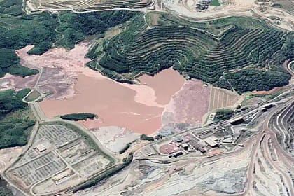 Imagem aérea mostra parte do Complexo Minerário de Itabira, com destaque para áreas de lavra, estruturas de beneficiamento e uma grande bacia com rejeitos de coloração avermelhada, cercada por taludes em formato de degraus. Ao redor, é possível observar vegetação, vias internas de acesso e instalações industriais ligadas à atividade mineradora. Registro relacionado às notícias de Itabira sobre projetos e operações no setor mineral.