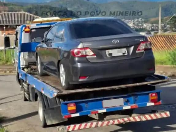 Um automóvel sedan de cor cinza escuro está sendo transportado sobre a plataforma de um caminhão-guincho azul, em via urbana. O veículo aparece visto pela traseira, com a placa parcialmente desfocada. Ao fundo, é possível observar casas, vegetação e uma área urbana em região montanhosa.