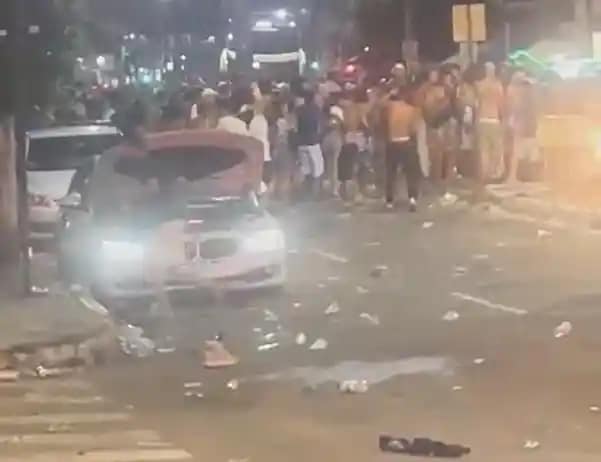 A imagem mostra uma avenida à noite, com grande concentração de pessoas ocupando a pista, com a presença da polícia em Monlevade. No primeiro plano, um carro branco aparece parado com os faróis acesos, cercado por lixo espalhado pelo asfalto, como copos e garrafas descartáveis. Ao fundo, é possível ver uma multidão reunida em frente aos veículos, ocupando os dois sentidos da via. A iluminação é típica de rua, com postes e luzes de estabelecimentos ao redor.
