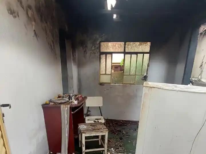 O interior de uma residência aparece severamente danificado por incêndio. As paredes e o teto estão cobertos por fuligem escura, com sinais evidentes de que o fogo se concentrou principalmente na parte superior do cômodo. Há uma abertura no telhado por onde entra luz natural. No ambiente, é possível ver móveis parcialmente queimados, como um pequeno armário, uma cadeira e uma mesa, além de objetos espalhados pelo chão.