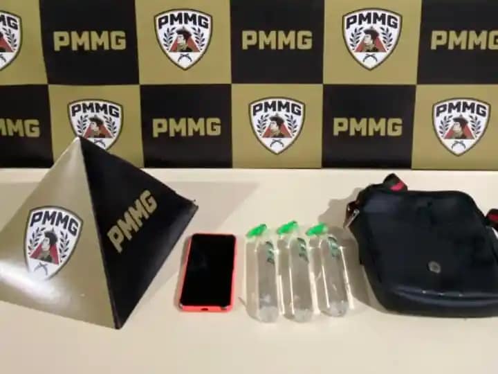 A imagem mostra os materiais apreendidos pela Polícia Militar durante a ocorrência registrada em Nova Era. Sobre uma mesa branca, estão expostos três frascos de lança-perfume, um aparelho celular com capa vermelha e uma bolsa preta tipo pochete. Ao lado dos objetos, aparece um suporte triangular com a sigla da corporação, posicionado diante de um painel ao fundo com o padrão xadrez nas cores preta e amarela, estampado com o emblema da PMMG. A foto integra as notícias de Nova Era relacionadas à prisão de dois suspeitos por tráfico de drogas durante evento carnavalesco no Centro da cidade.