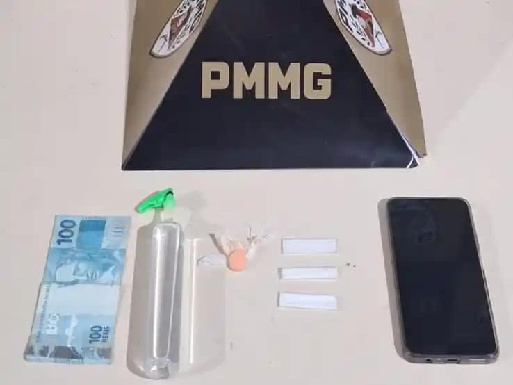 A imagem mostra materiais apreendidos pela Polícia Militar, dispostos sobre uma superfície clara, com um suporte ao fundo exibindo a sigla “PMMG”. Entre os itens estão uma cédula de R$ 100, um frasco plástico contendo lança-perfume, três papelotes de substância semelhante à cocaína, um comprimido de ecstasy e um aparelho celular. Os objetos estão organizados lado a lado, compondo o registro da ocorrência policial. Notícias de nova era