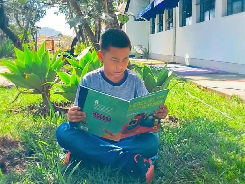 A imagem mostra uma criança sentada sobre a grama, em um ambiente externo e arborizado, lendo um livro infantil. Ela está sentada com as pernas cruzadas, concentrada na leitura, segurando um livro de capa verde com ilustrações coloridas.