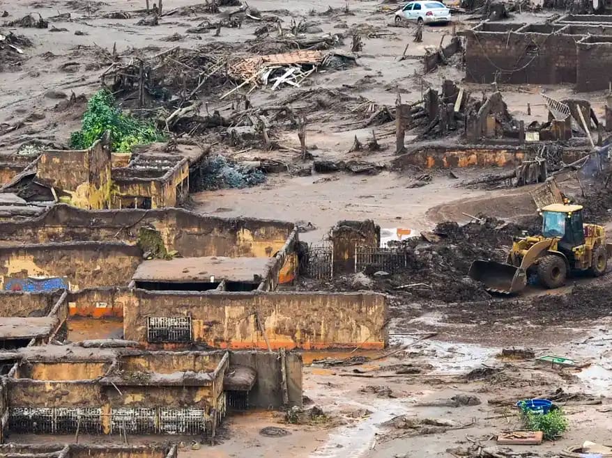 Imagem aérea mostra área devastada pelo rompimento da barragem em Brumadinho. Construções aparecem parcialmente soterradas pela lama, com paredes destruídas e estruturas expostas. O terreno está coberto por rejeitos de mineração, entulho e troncos de árvores.