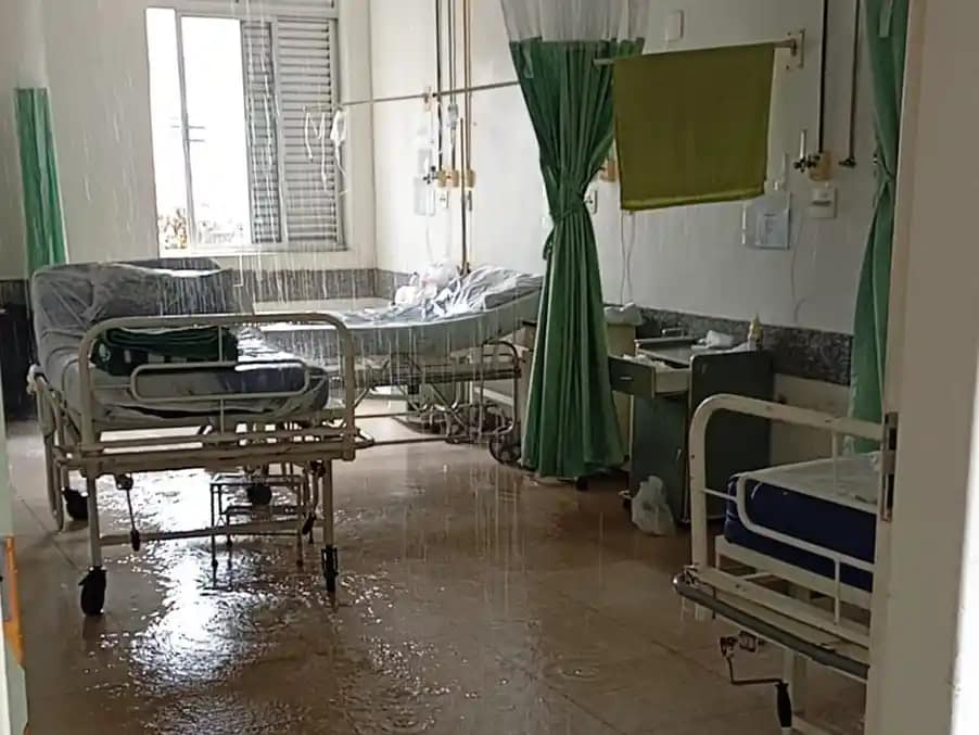 Hospital em João Monlevade com enfermaria masculina parcialmente alagada após vazamento no teto. A imagem mostra leitos hospitalares posicionados lado a lado, com água escorrendo do forro e formando poças no piso. Parte do teto apresenta infiltração visível, e o chão está molhado em diversos pontos, refletindo a iluminação do ambiente.