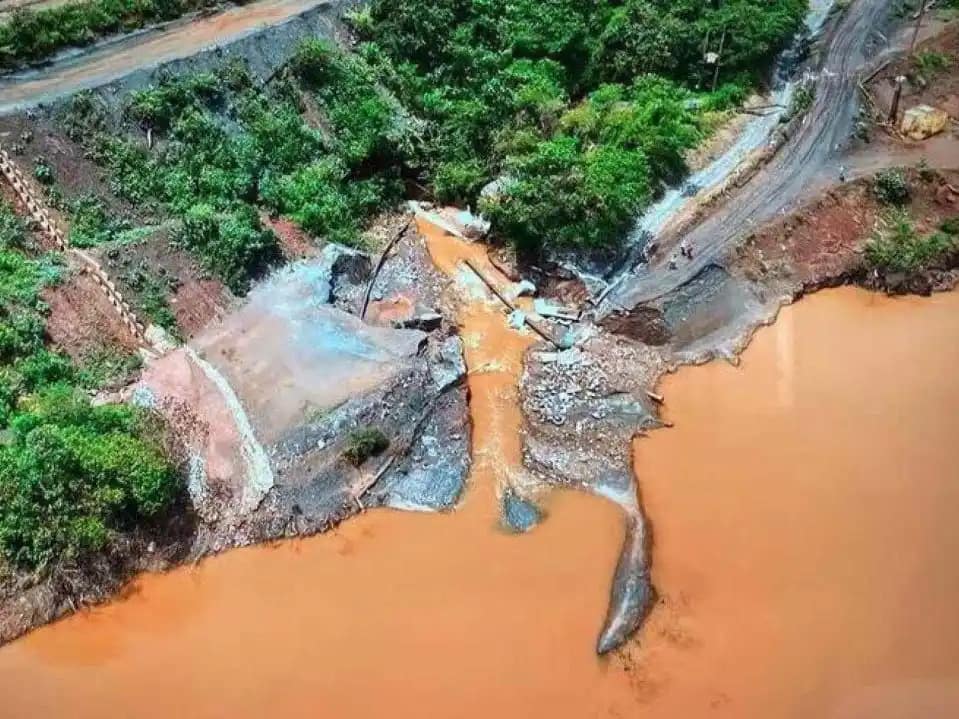 Imagem aérea mostra área afetada por rompimento de estrutura de contenção, com grande volume de lama alaranjada avançando por um vale, estradas danificadas, vegetação ao redor e trechos de terra expostos, evidenciando impacto ambiental e erosão do terreno.