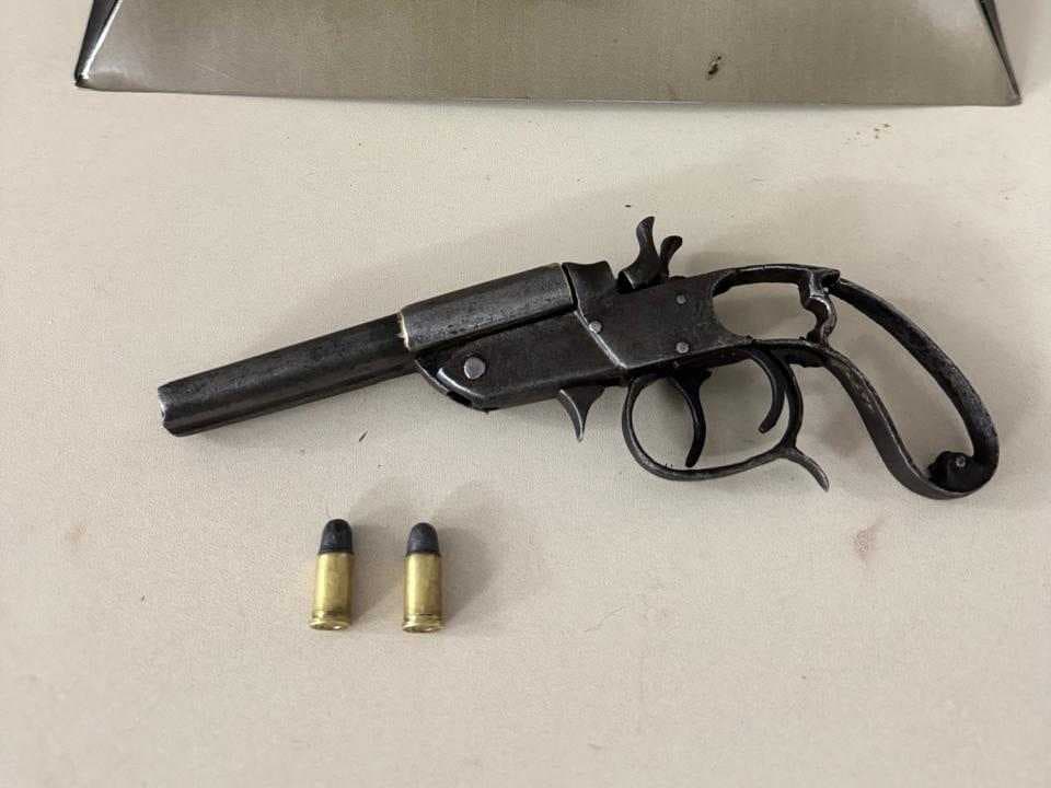 Imagem mostra uma arma de fogo do tipo garrucha apoiada sobre uma superfície clara, acompanhada de duas munições intactas posicionadas à frente. Ao fundo, aparece um suporte metálico em formato triangular com o emblema da Polícia Militar, indicando que o material foi apreendido e apresentado em ambiente institucional. A cena sugere registro oficial de apreensão realizada pelas forças de segurança.