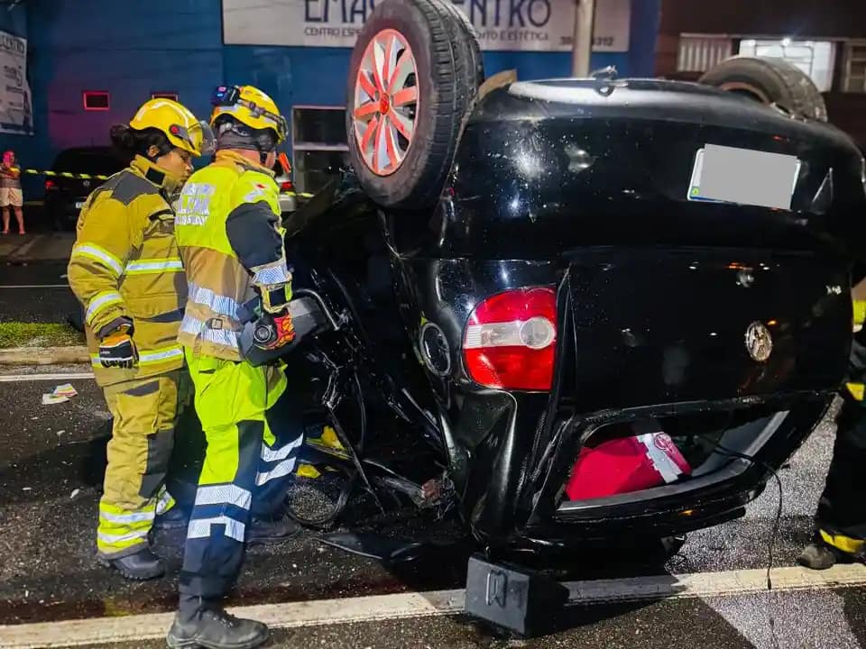 A imagem mostra um veículo preto capotado após acidente em Monlevade, com as rodas voltadas para cima, imobilizado no meio de uma via urbana durante a noite. O automóvel apresenta danos significativos na lataria, especialmente na parte traseira e lateral, indicando a força do impacto. Ao lado do carro, dois militares do Corpo de Bombeiros, devidamente equipados com capacetes, lanternas e uniformes de proteção, atuam no atendimento à ocorrência.